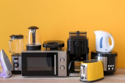 Különböző háztartási eszközök, mint például vízforraló, kávéfőző és mikrohullámú sütő egy sárga háttér előtt.