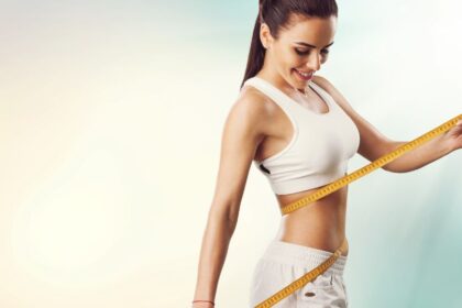 Egy fiatal nő mérőszalaggal méri a derekát, mosolyogva áll a kamera előtt.