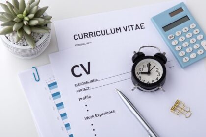 Egy önéletrajz papír, óra, toll és számológép látható egy asztalon, zöld növénnyel.