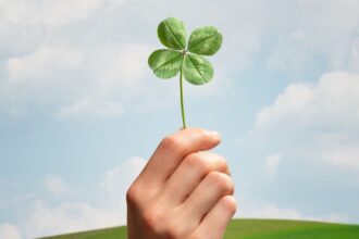 Egy kéz tart egy négylevelű lóherét a kék ég alatt, szimbolizálva a szerencsét és a spiritualitást.