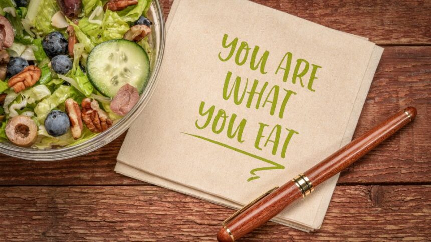 Friss saláta tál, tele zöldségekkel és gyümölcsökkel, mellett egy papíralátét, amelyen a 'You are what you eat' felirat olvasható.