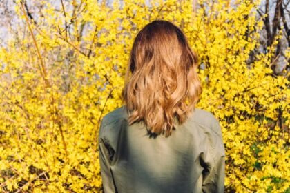 Egy nő hátulról, hullámos hosszú bob frizurával, sárga virágokkal teli háttér előtt.