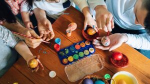 Családtagok közösen festenek húsvéti tojásokat, színes festékek és ecsetek mellett.