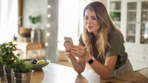 Egy fiatal nő a konyhában ülve nézi a telefonját, miközben a háttérben zöld növények és gyümölcsök láthatók.