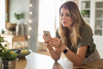 Egy fiatal nő a konyhában ülve nézi a telefonját, miközben a háttérben zöld növények és gyümölcsök láthatók.