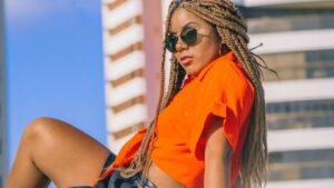 womensway 128 Egy fiatal nő narancssárga blúzban és napszemüvegben, modern városi háttér előtt.