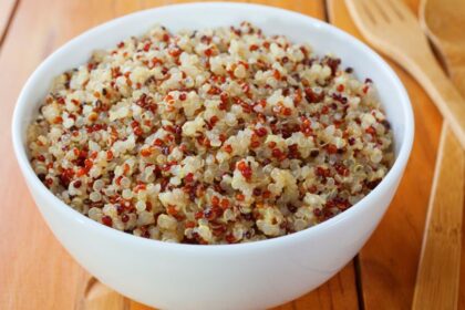 Egy fehér tálban főtt quinoa látható, különböző színű szemekkel, fából készült evőeszközök mellett.