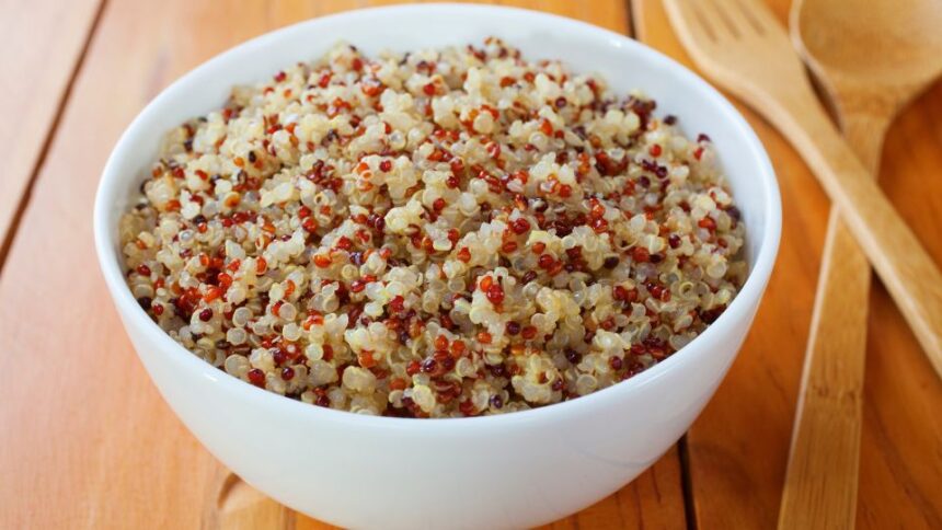 Egy fehér tálban főtt quinoa látható, különböző színű szemekkel, fából készült evőeszközök mellett.