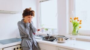 Egy fáradt nő áll a konyhában, kezét az arcán tartva, miközben a mosogató mellett áll.