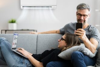 Egy pár a kanapén ül, a légkondicionáló hűs levegője alatt, laptopot és okostelefont használva.