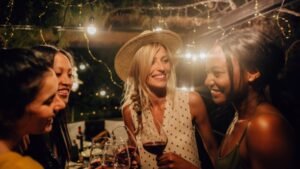 womensway 133 Öt barátnő vidáman beszélget egy hangulatos teraszon, poharaikban borral, a háttérben fények ragyognak.