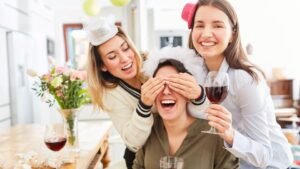 womensway 131 Három nő vidáman ünnepel otthon, az egyikük szemét eltakarják, míg a másikok pohár borral a kezükben nevetnek.