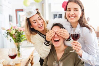 Három nő vidáman ünnepel otthon, az egyikük szemét eltakarják, míg a másikok pohár borral a kezükben nevetnek.