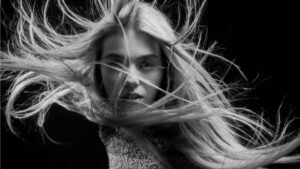Egy fiatal nő arca látható, hosszú, szőke haja szélben lobog, a háttér fekete.