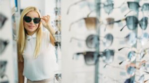 Egy fiatal nő napszemüveget próbál egy boltban, mögötte különböző keretek sorakoznak.