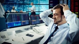 Egy frusztrált férfi ül egy tőzsdei irodában, számítógépek előtt, ahol piros számok és grafikonok láthatók.