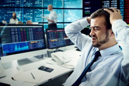Egy frusztrált férfi ül egy tőzsdei irodában, számítógépek előtt, ahol piros számok és grafikonok láthatók.