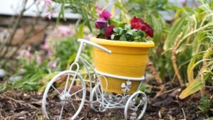 womensway 137 Egy sárga virágcserép egy fehér fém kerékpáron, színes virágokkal a kertben.