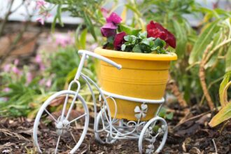 Egy sárga virágcserép egy fehér fém kerékpáron, színes virágokkal a kertben.