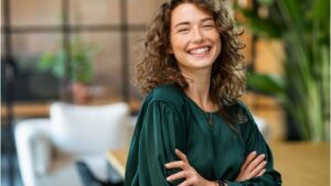 Egy magabiztos nő mosolyogva áll, karjait keresztbe fonva, zöld blúzban, modern környezetben.