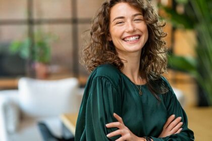 Egy magabiztos nő mosolyogva áll, karjait keresztbe fonva, zöld blúzban, modern környezetben.
