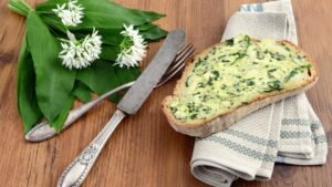 Friss medvehagyma levelek és virágok mellett egy medvehagymás kenyérszelet látható.