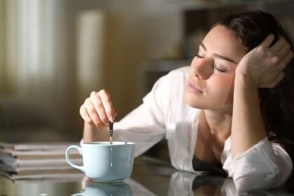 Egy fiatal nő álmosan ül az asztalnál, miközben egy csésze kávét kever.