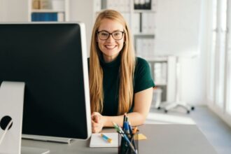 Egy fiatal nő mosolyogva ül az íróasztalánál, háttérben modern munkahelyi környezet.