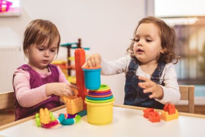 Két kisgyermek játszik színes építőkockákkal egy világos szobában.