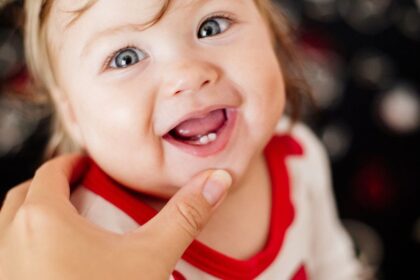 Egy boldog kisbaba mosolyog, látható az első foga, miközben egy felnőtt kéz simogatja az állát.