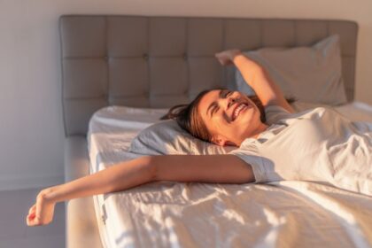 Egy fiatal nő boldogan ébred a napfényben, karjait kitárva fekszik az ágyban.
