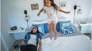 Egy kislány ugrál az ágyon, miközben édesanyja nevetve fekszik mellette.