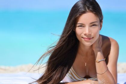 Egy fiatal nő napozik a tengerparton, hosszú hajjal és vidám arccal.