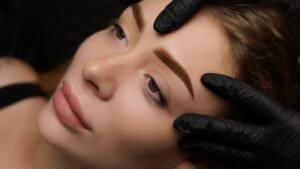 Egy fiatal nő arca, sminkkel, miközben egy szakember kezével formázza a szemöldökét.