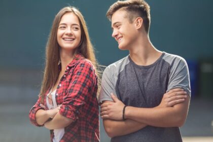 Két fiatal, egy lány és egy fiú, barátságosan mosolyognak egymásra, miközben a háttérben zöld szemetesek láthatók.