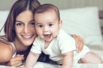 Egy boldog anya és kisbabája együtt fekszenek az ágyon, mosolyogva néznek a kamerába.