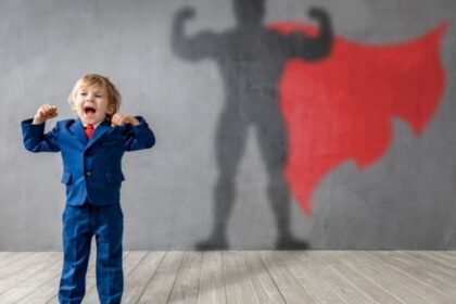 Egy kisfiú kék öltönyben áll, karjait felemelve, miközben szuperhős árnyéka látható a falon.