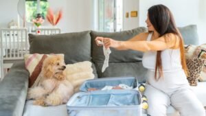 Egy kismama a kanapén ülve babaruhákat pakol egy bőröndbe, mellette egy kutyával.