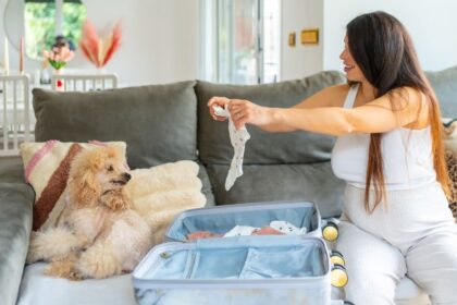 Egy kismama a kanapén ülve babaruhákat pakol egy bőröndbe, mellette egy kutyával.