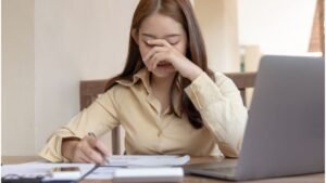 womensway 150 Egy fiatal nő az asztalnál ül, kezével az arcát takarja, láthatóan stresszes vagy szomorú, miközben jegyzetel.