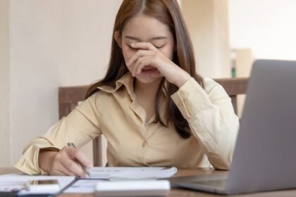 Egy fiatal nő az asztalnál ül, kezével az arcát takarja, láthatóan stresszes vagy szomorú, miközben jegyzetel.