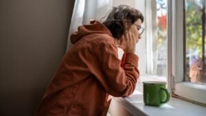 womensway 148 Egy introvertált nő az ablak mellett ül, gondolkodva néz kifelé, kezében egy zöld bögre.