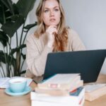 Egy fiatal nő gondolkodik a laptopja előtt, könyvek és egy csésze kávé mellett ülve.