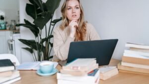 Egy fiatal nő gondolkodik a laptopja előtt, könyvek és egy csésze kávé mellett ülve.