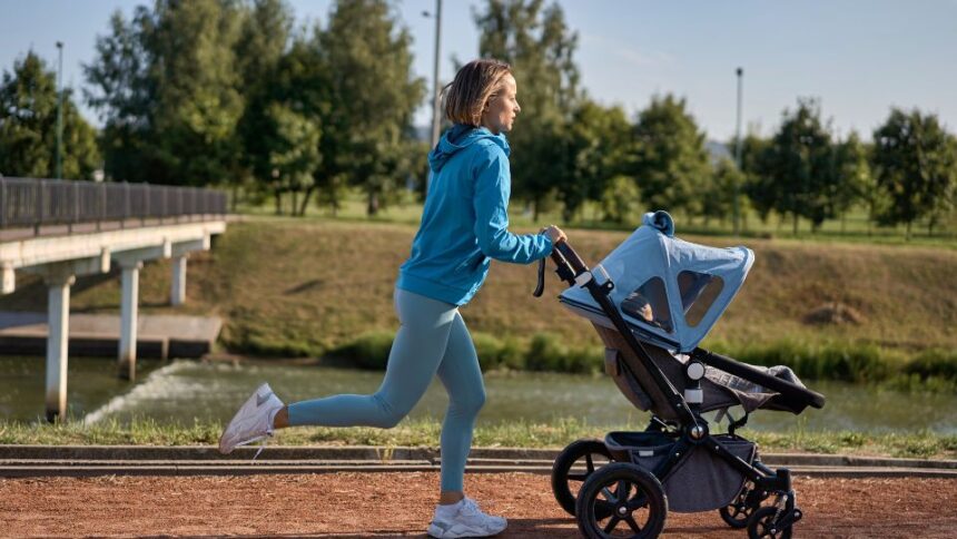 Egy fiatal nő babakocsit tolva fut a parkban, napfényben, zöld fák között.