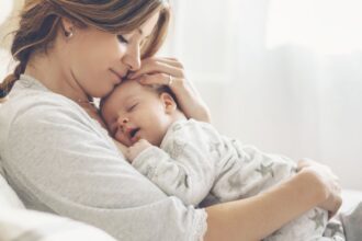 Egy édesanya ölelésében tartja alvó babáját, a napfény lágyan besüt a szobába.