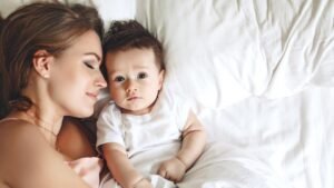 womensway 140 Egy édesanya és újszülött babája fekszik az ágyon, az anya mosolyogva néz a babára, aki kíváncsian figyel.