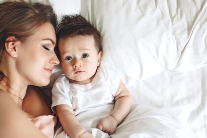 Egy édesanya és újszülött babája fekszik az ágyon, az anya mosolyogva néz a babára, aki kíváncsian figyel.