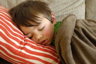 Egy kisfiú békésen alszik egy piros csíkos párnán, takaróba burkolózva.