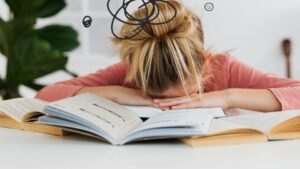 Egy fiatal nő könyvek között ül, kezét a fejére téve, a halogatás és stressz jeleit mutatja.